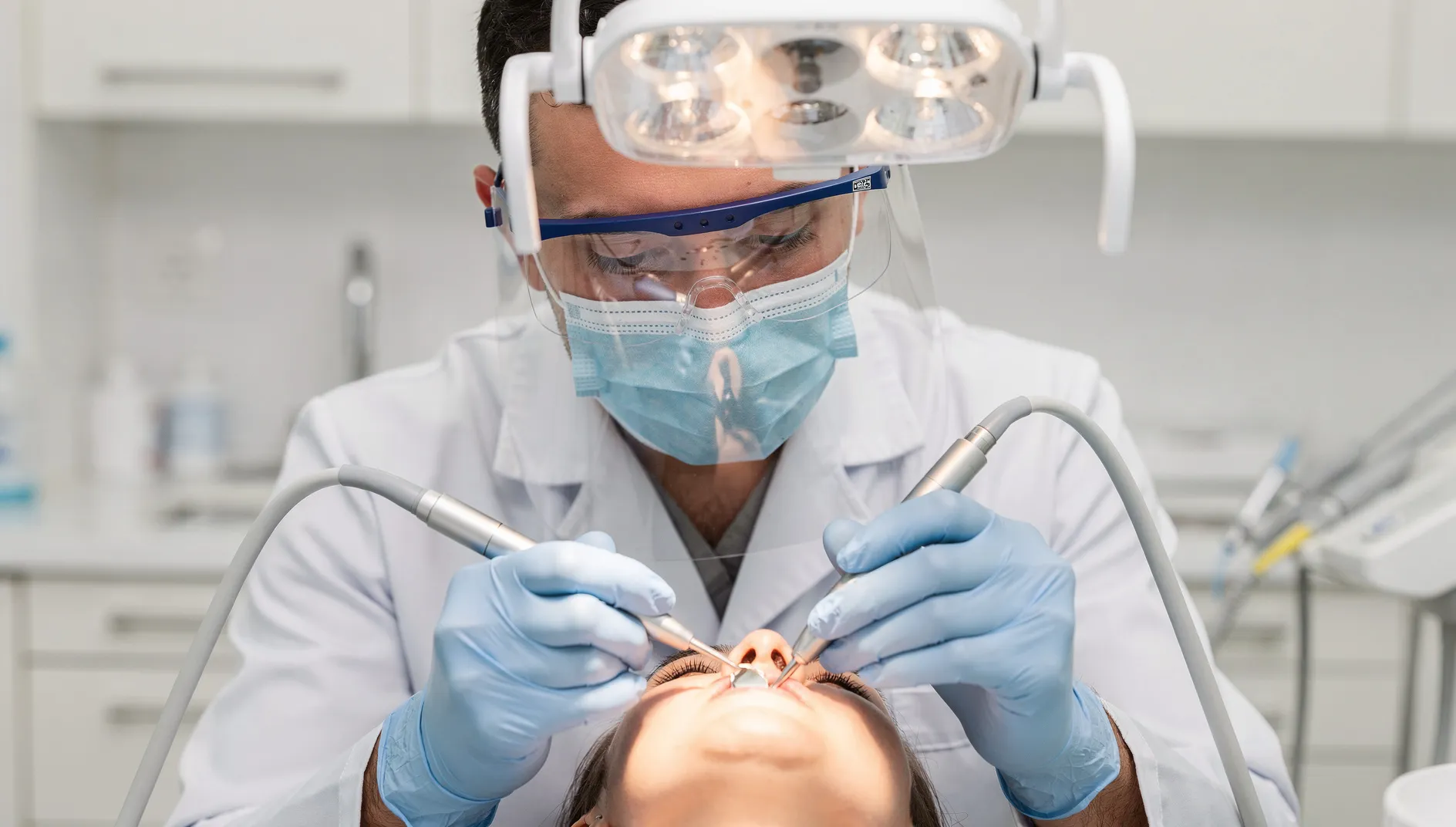 A dentist in a mask and face shield works on a patient with two instruments under the operating light
